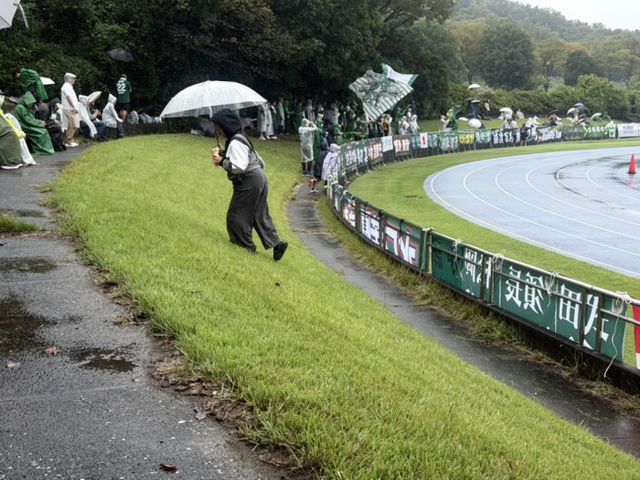 雨の中、傘やレインコート姿のサポーターが並ぶゴール裏の芝生席